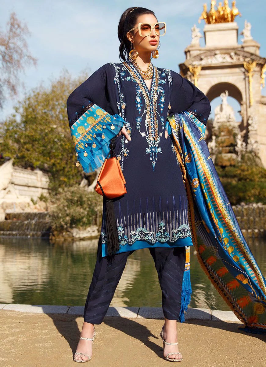 A woman models a traditional South Asian outfit featuring a navy blue kurta with intricate gold and blue embroidery, matching navy trousers, and a vibrant blue dupatta with gold and orange patterns. She is standing outdoors near a body of water with a historic stone archway in the background.