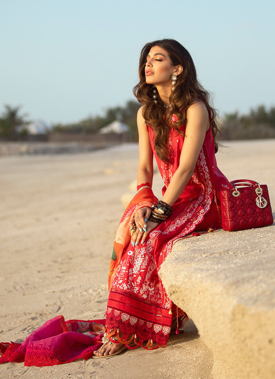 A woman sits on a stone wall in a sandy outdoor setting, wearing a bright red and orange printed traditional dress with a matching dupatta. A red quilted handbag rests beside her.