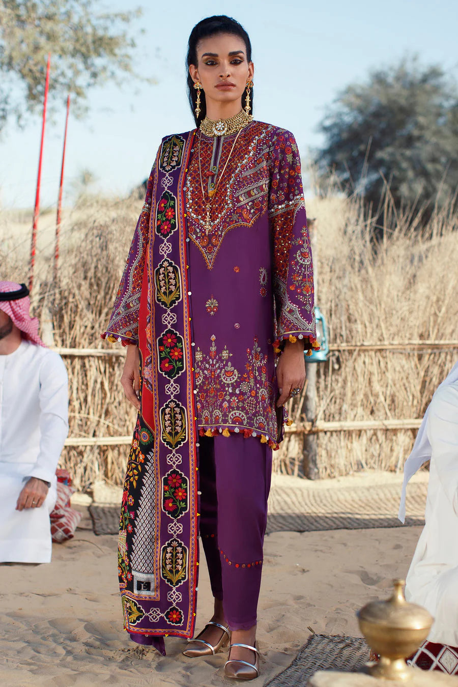 A woman stands outdoors in a desert-like setting wearing a traditional purple three-piece outfit with ornate multicolor embroidery and a matching patterned dupatta. Two men in white traditional clothing are partially visible at the left and right edges.