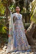 A woman stands outdoors among trees and rocks wearing a long printed dress with a matching sheer dupatta. The outfit has a light base with blue, orange, and red floral and botanical patterns.