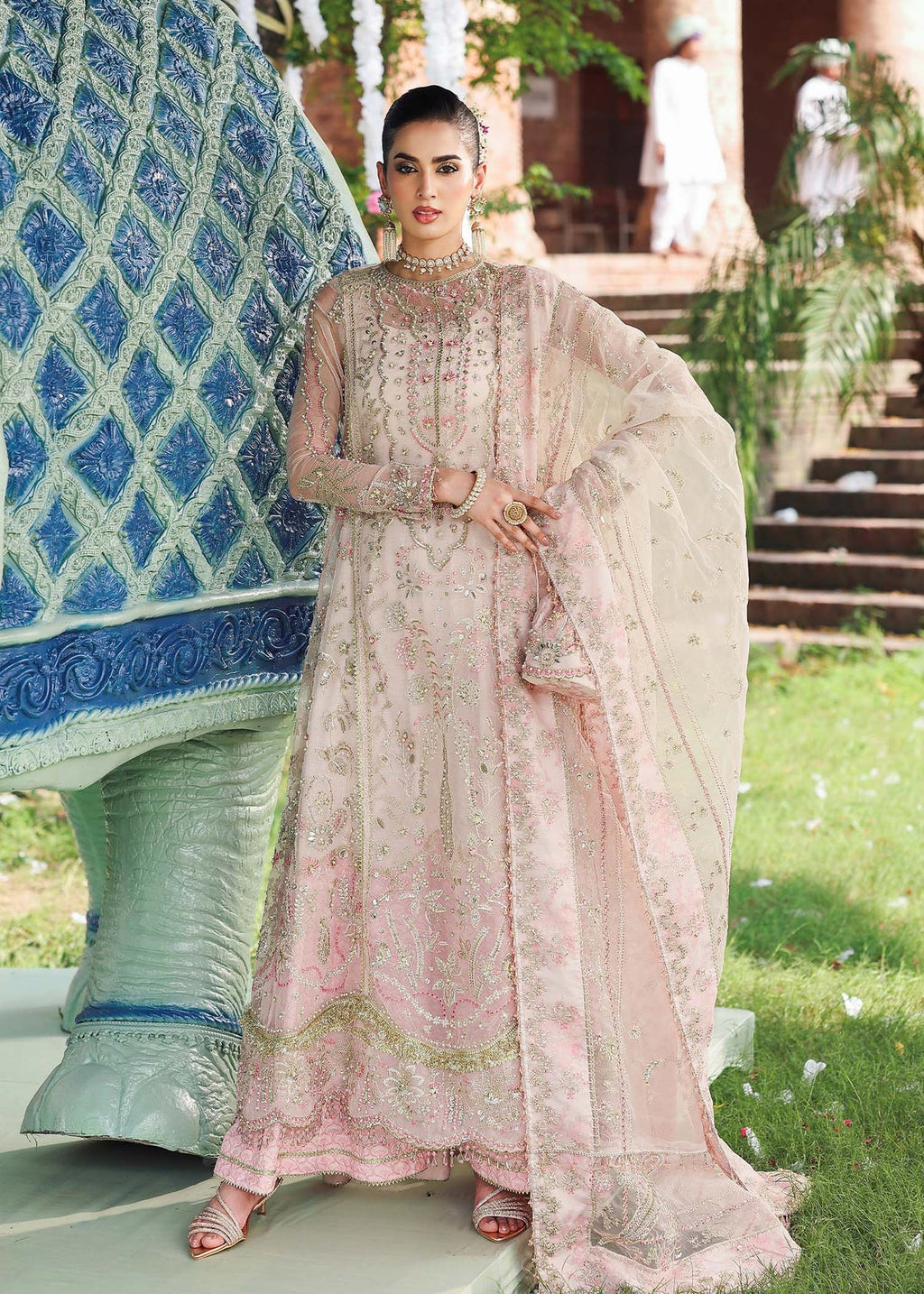 Woman in a traditional embroidered outfit standing outdoors with greenery and architectural elements in the background.