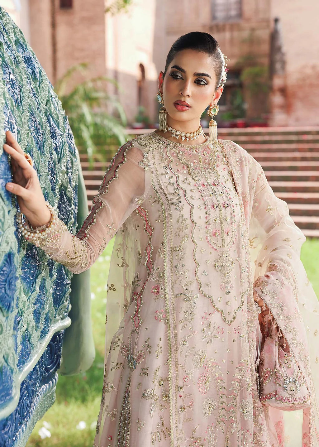 Woman in a traditional embroidered outfit standing outdoors with a decorative wall and plants in the background.