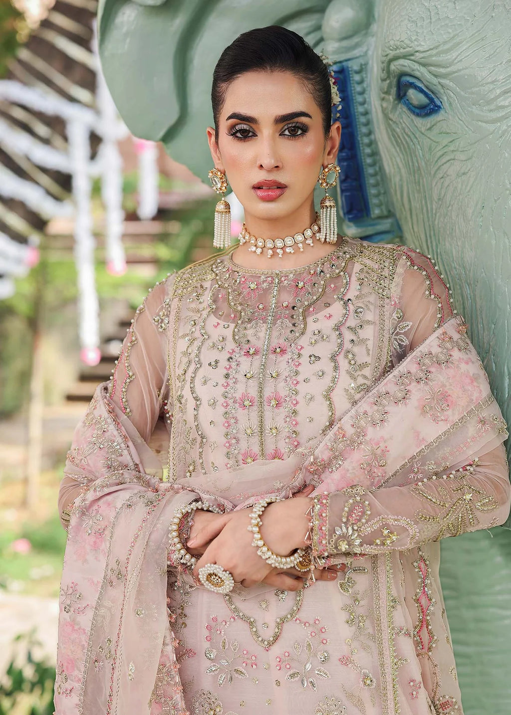 Woman in traditional pink embroidered outfit with jewelry, standing in front of an elephant statue.