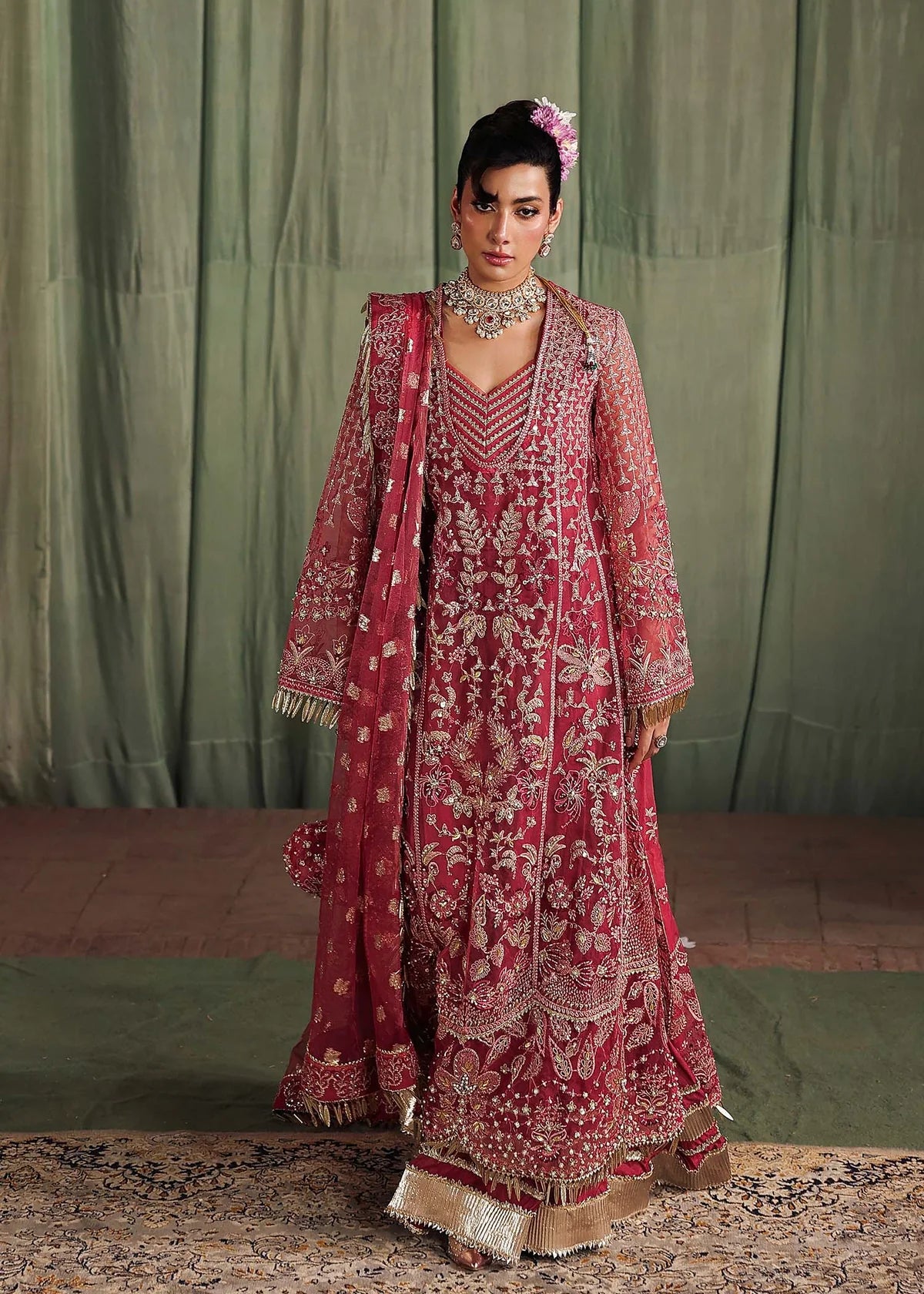Woman in a traditional maroon and gold embroidered outfit with a matching shawl against a green curtain background.