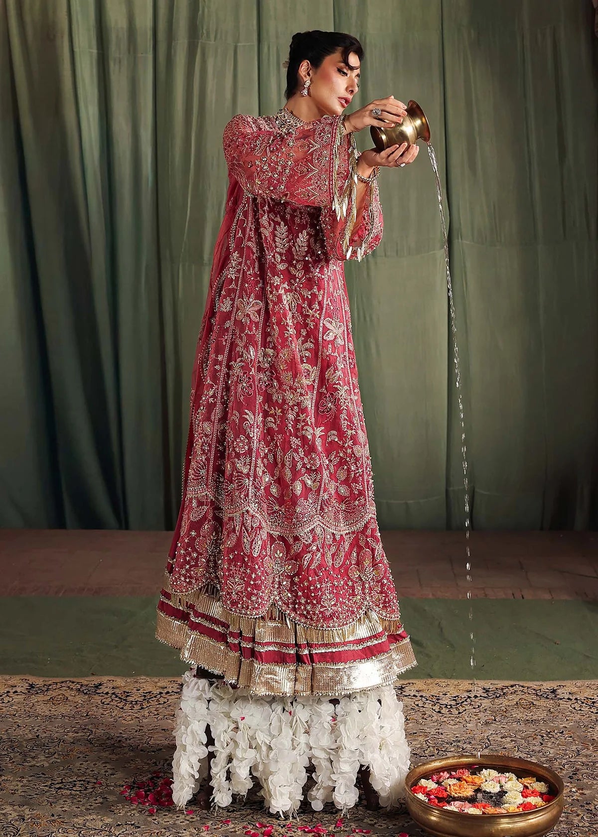 Woman in a red and gold traditional outfit standing on a floral stool against a green curtain.