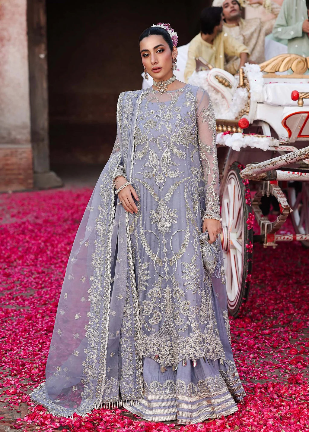 Woman in a gray embroidered traditional outfit standing on red petals with a decorated cart in the background.