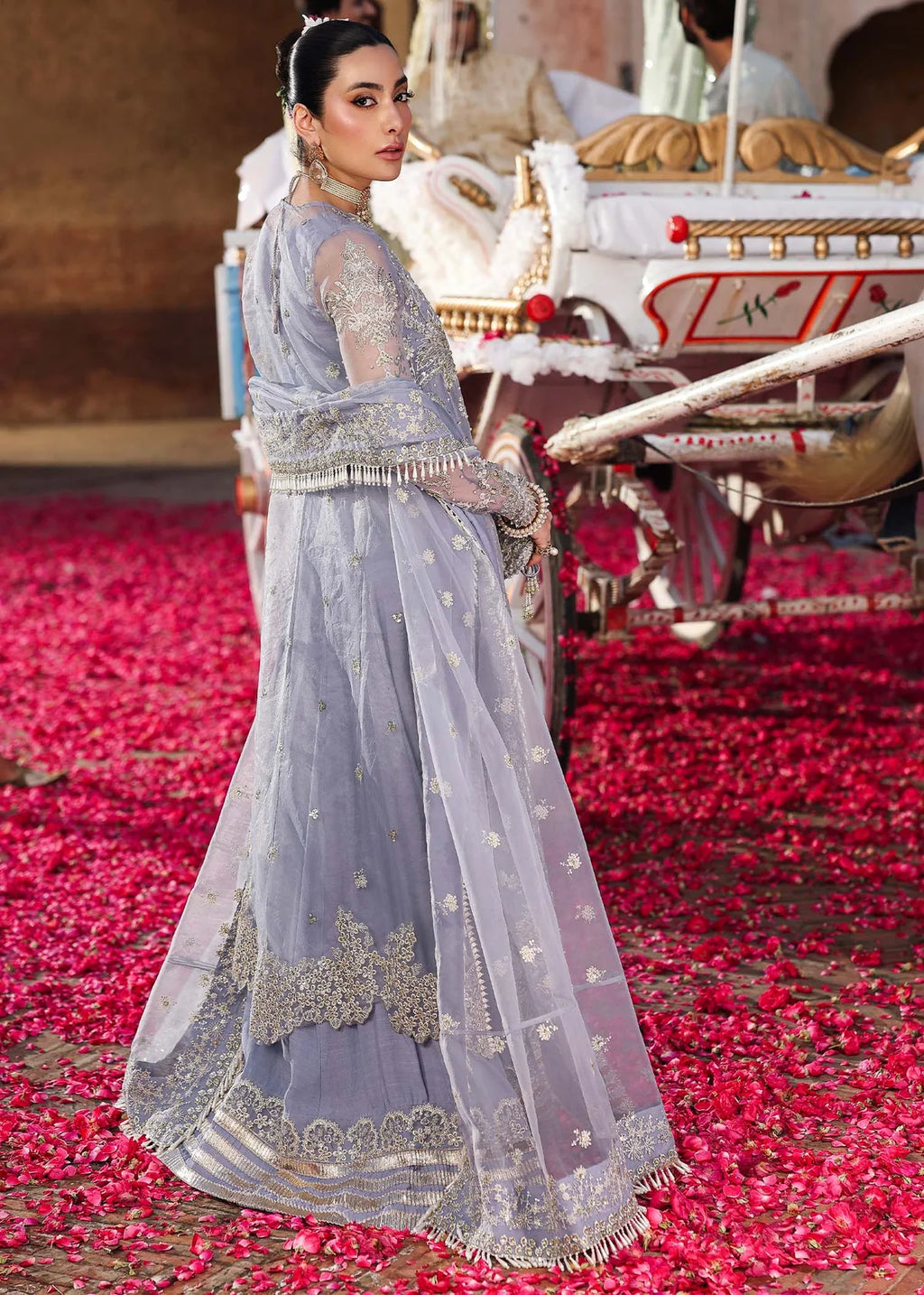 Woman in a traditional outfit standing on a path with red flowers, with a vehicle in the background.