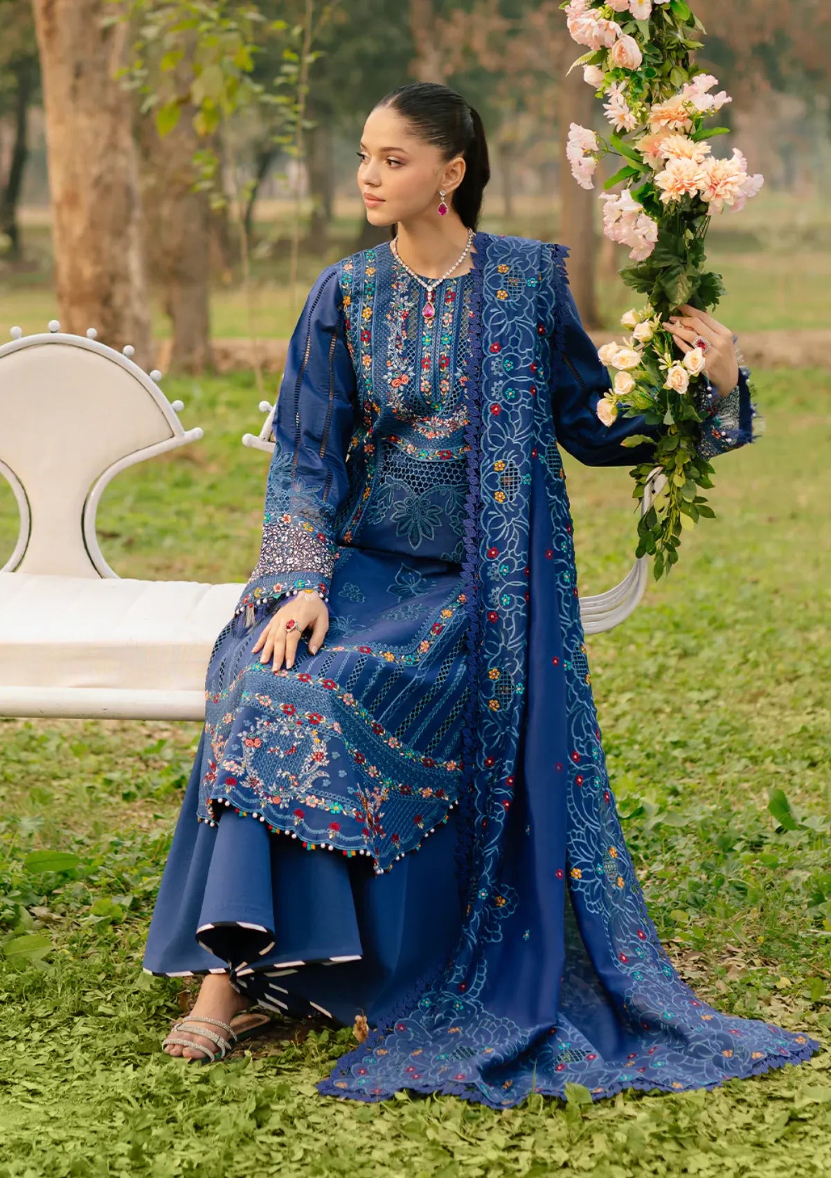 A woman seated outdoors on a white bench wearing a rich royal blue traditional outfit with intricate multicolor embroidery and a matching draped scarf. She is holding a floral garland in a grassy park setting.
