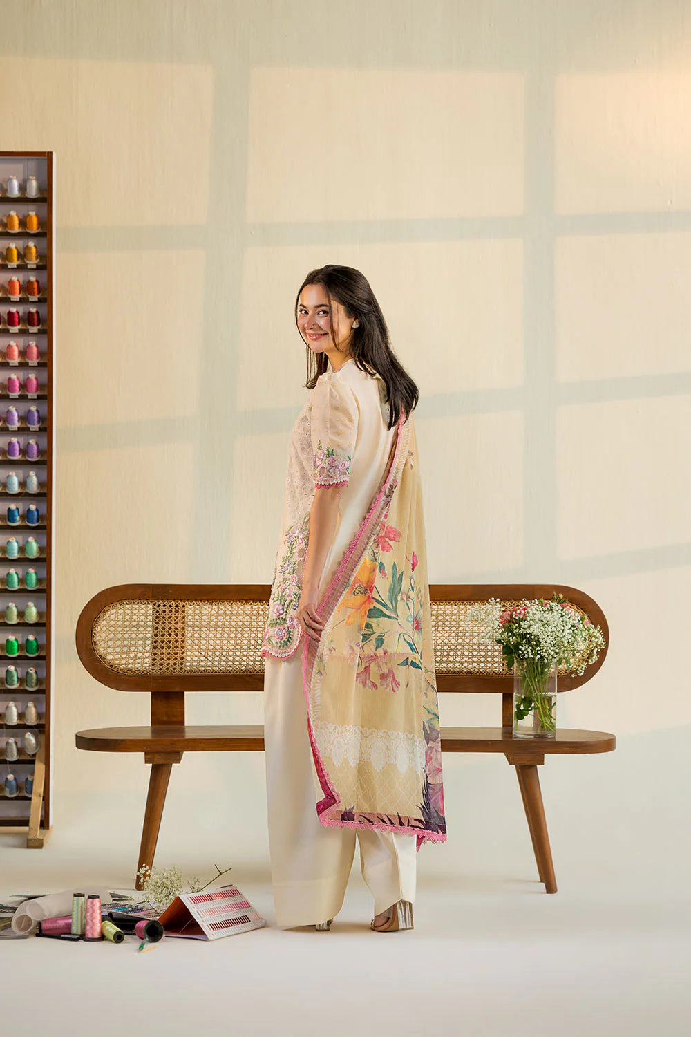 Woman in a floral outfit standing next to a wooden bench with a vase of flowers in a minimalistic room.