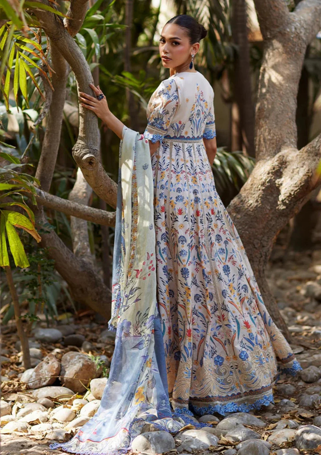 A woman stands outdoors among trees and rocks wearing a long printed dress with a matching dupatta. The dress has a light beige base with blue, orange, and green floral and paisley-style print details.