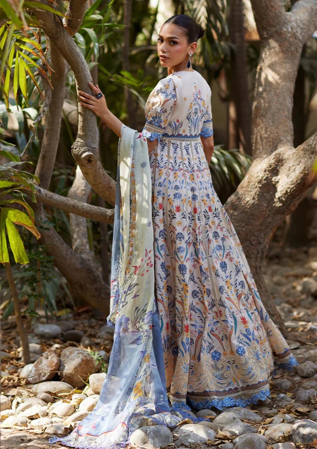 A woman stands outdoors among trees and rocks wearing a long printed dress with a matching dupatta. The dress has a light beige base with blue, orange, and green floral and paisley-style print details.
