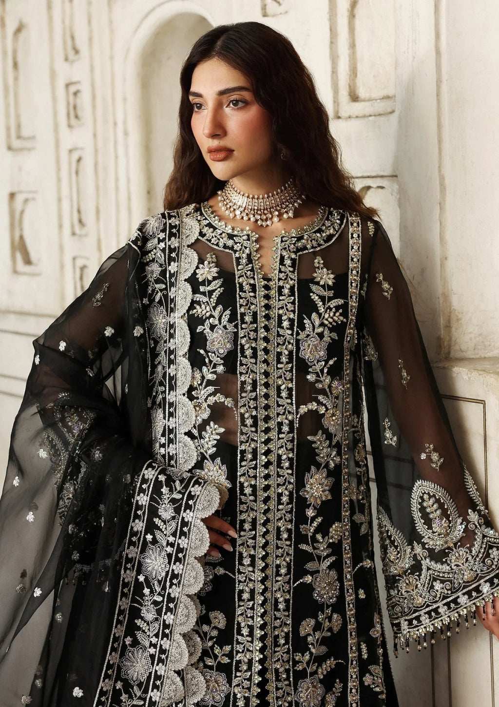 A woman wearing a black embroidered traditional outfit with sheer sleeves and ornate silver floral detailing, posed against a light stone wall.