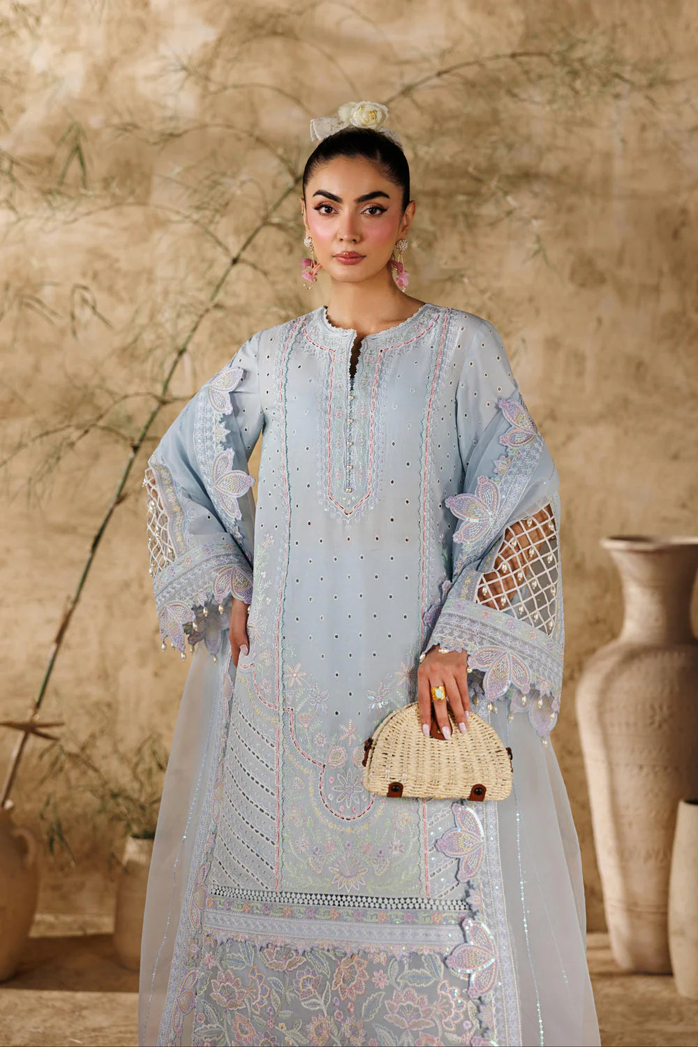 A woman models a light blue embroidered traditional long dress with sheer sleeves and intricate lace detailing, holding a woven beige clutch against a textured beige background.