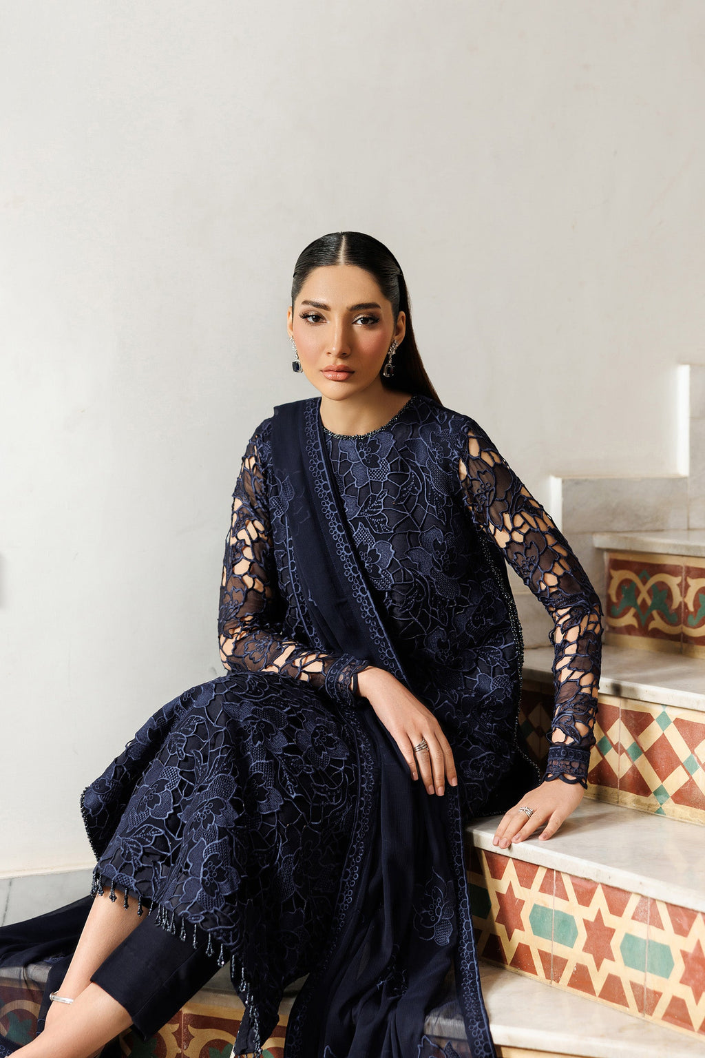Woman in a dark blue lace outfit sitting on a patterned stool against a white wall.
