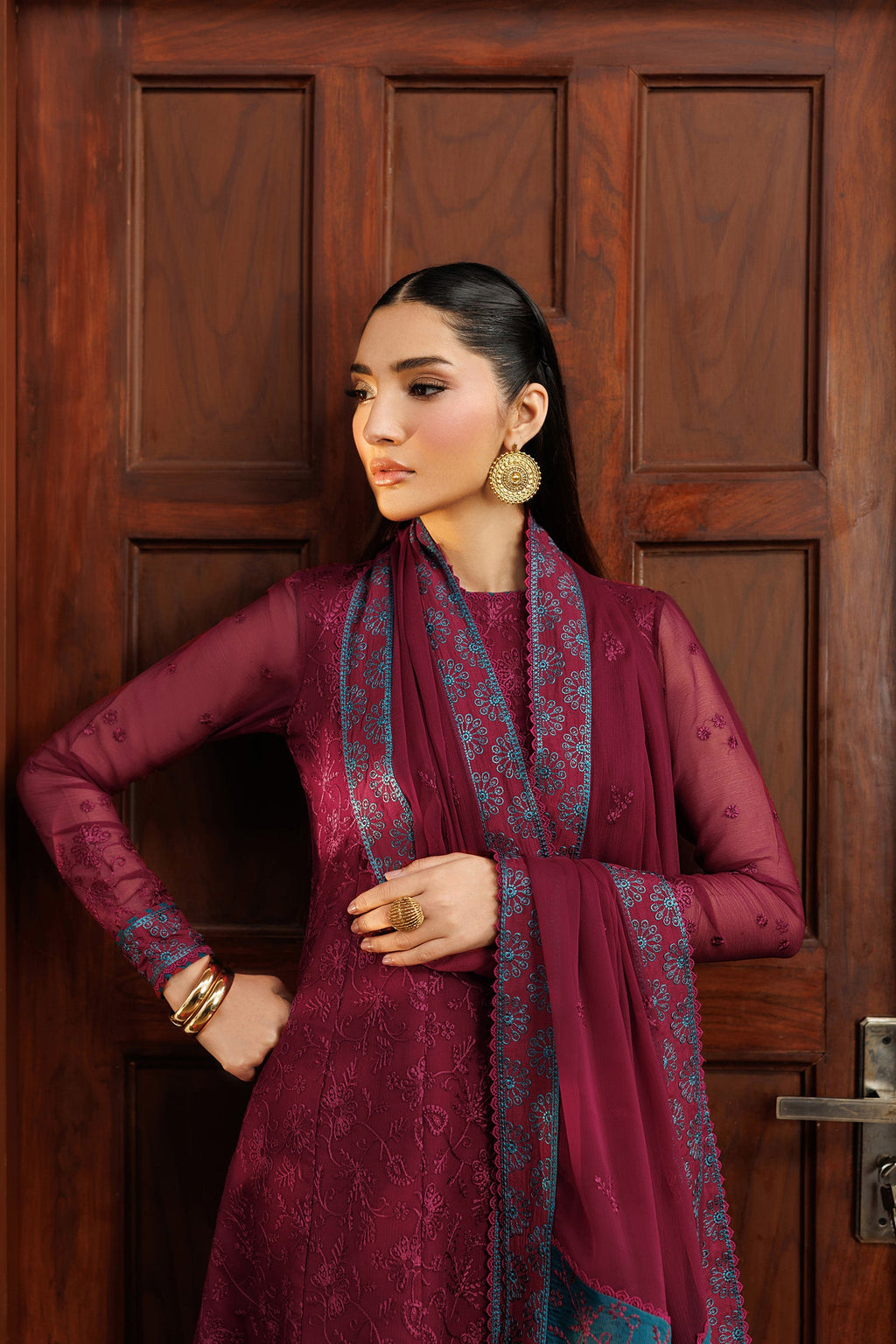 Woman in a maroon traditional outfit with a patterned scarf standing against a wooden door.