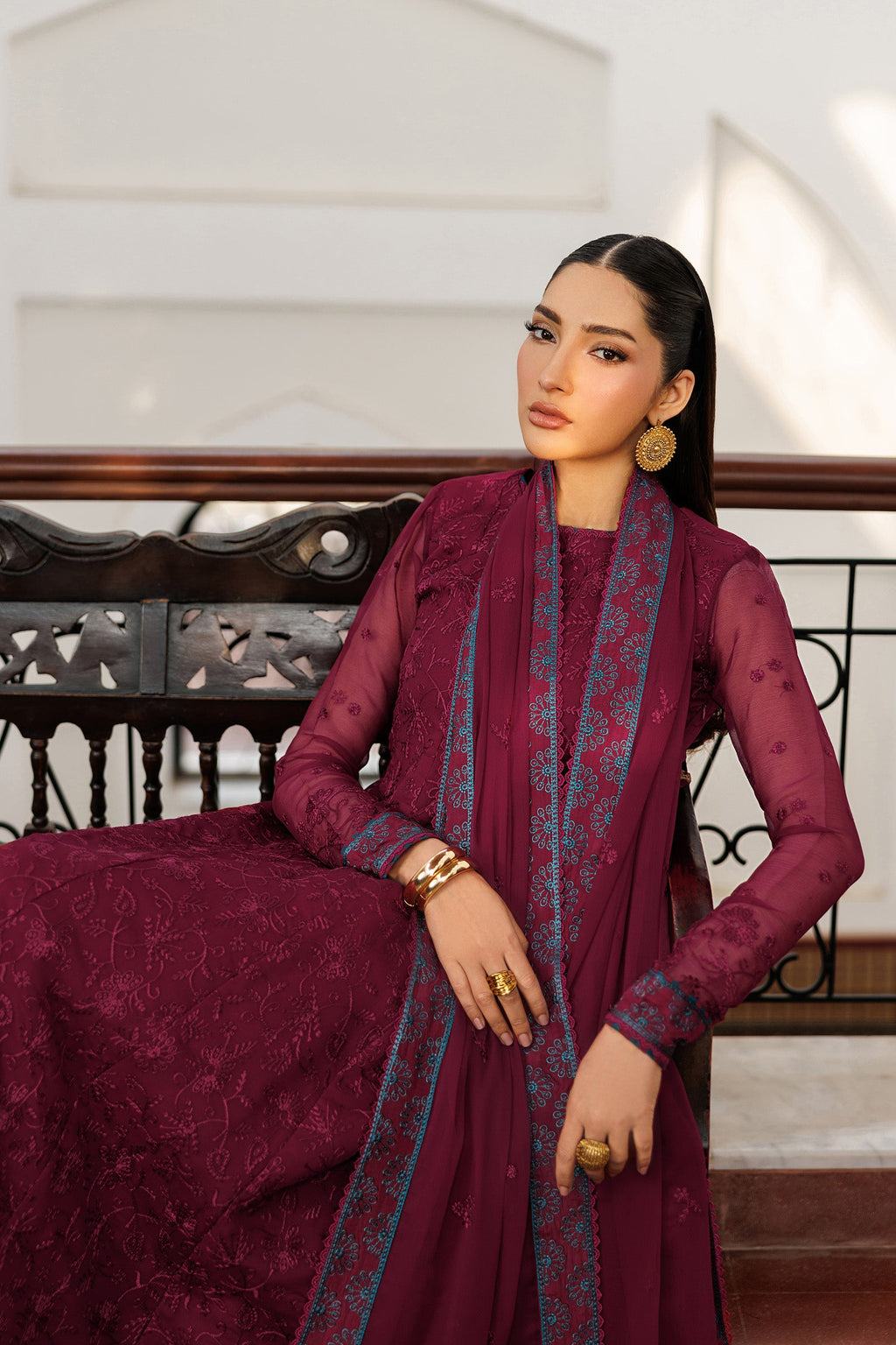 Woman in a maroon traditional outfit with intricate patterns sitting on a decorative bench.