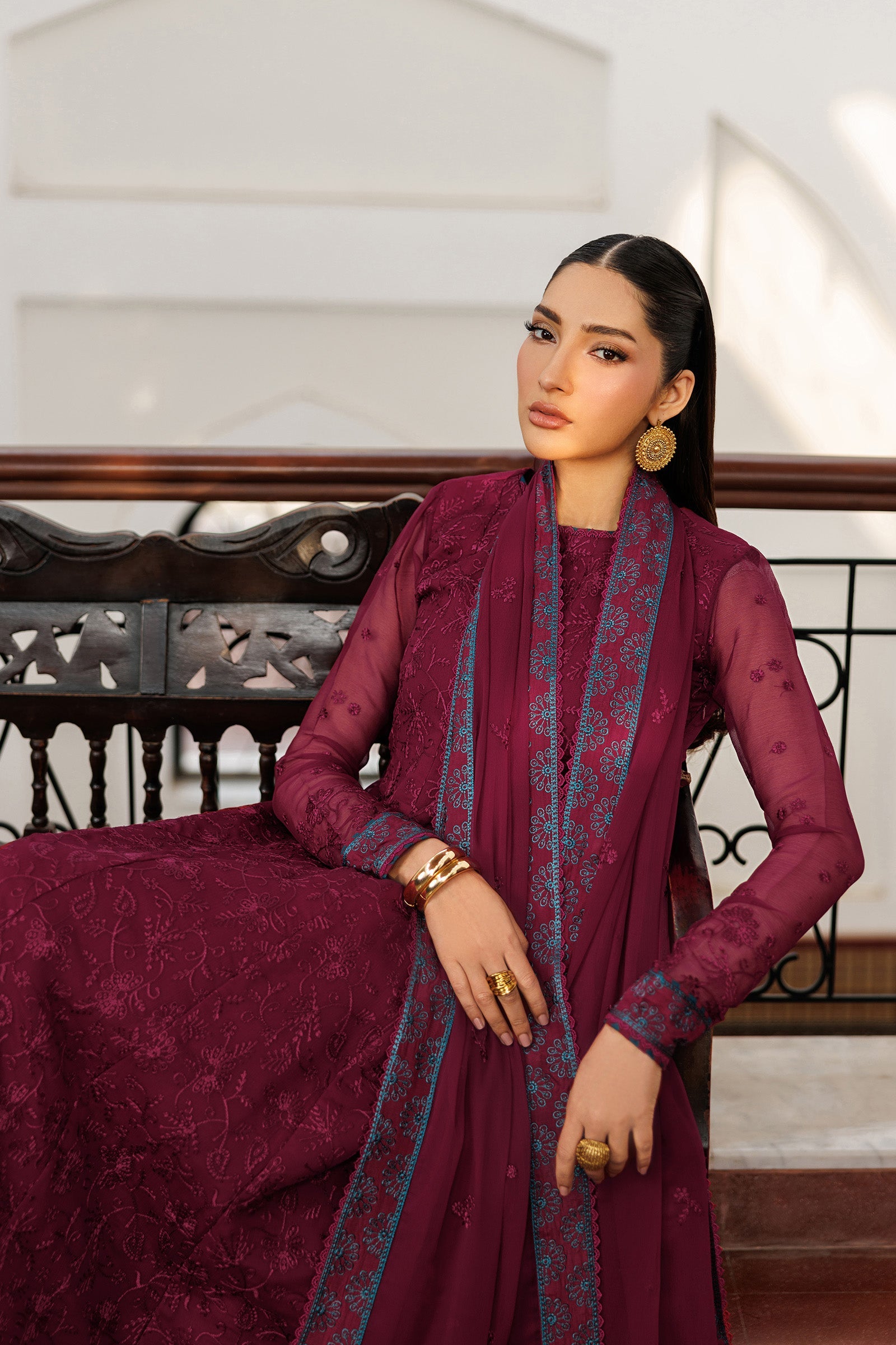 Woman in a maroon traditional outfit with intricate patterns sitting on a decorative bench.