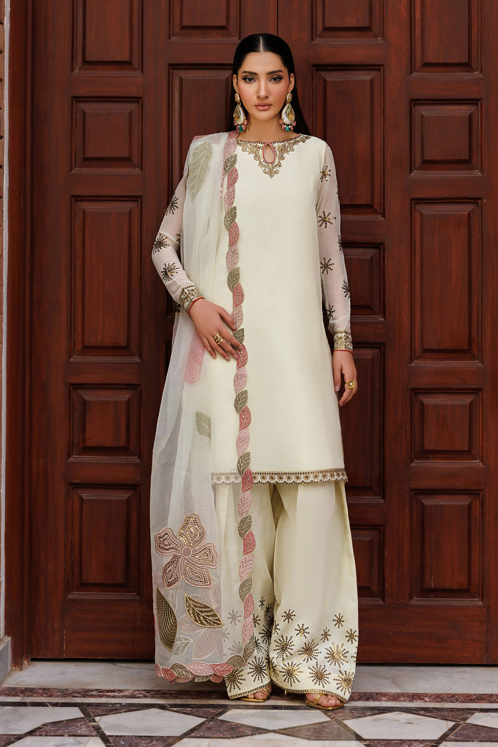 Woman in traditional white outfit with floral patterns standing in front of a wooden door.