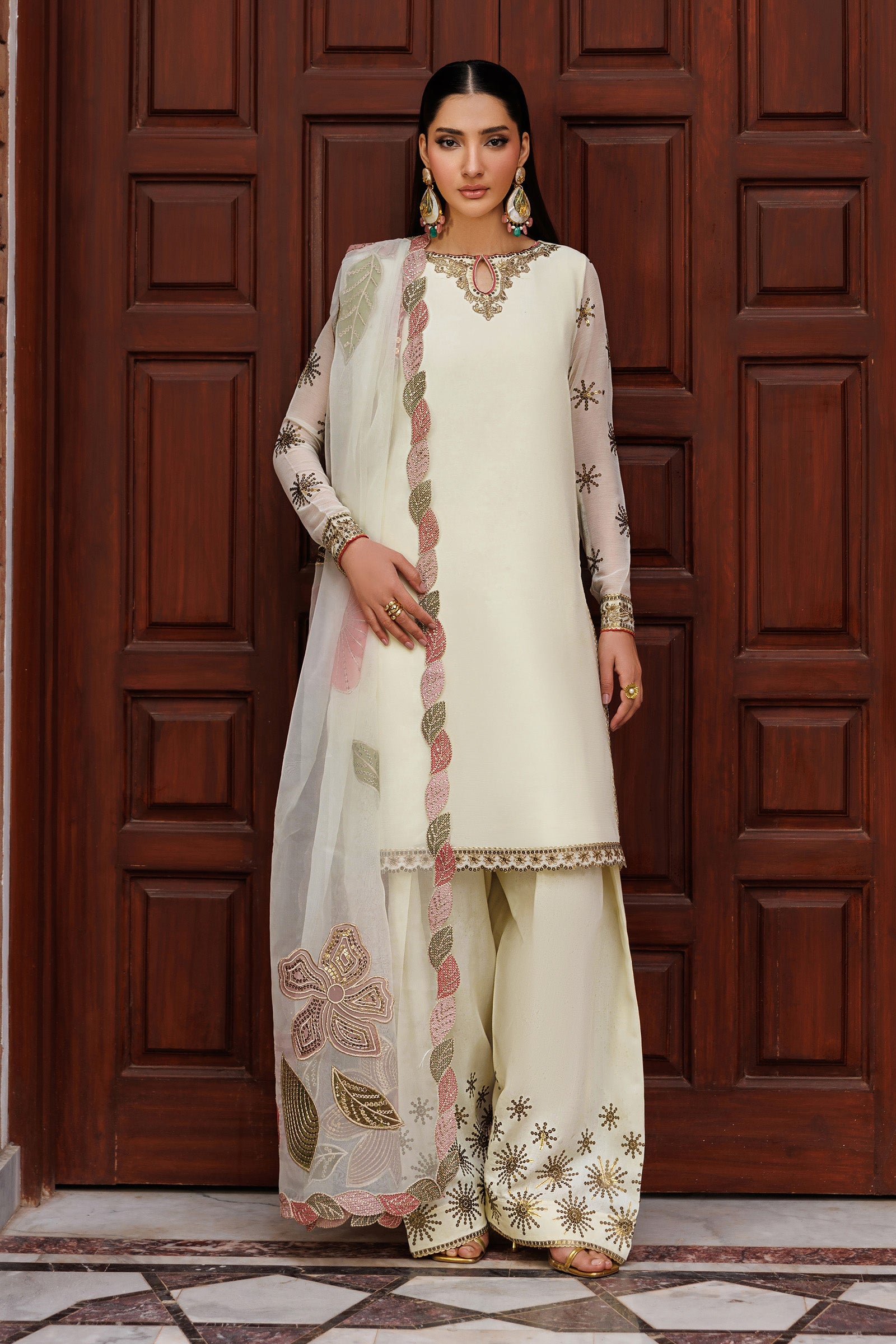 Woman in traditional white outfit with floral patterns standing in front of a wooden door.