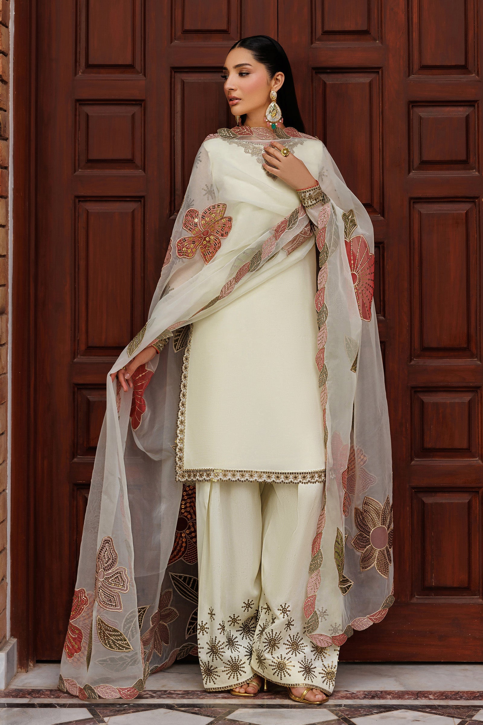 Woman wearing a traditional white outfit with floral patterns in front of a wooden door.