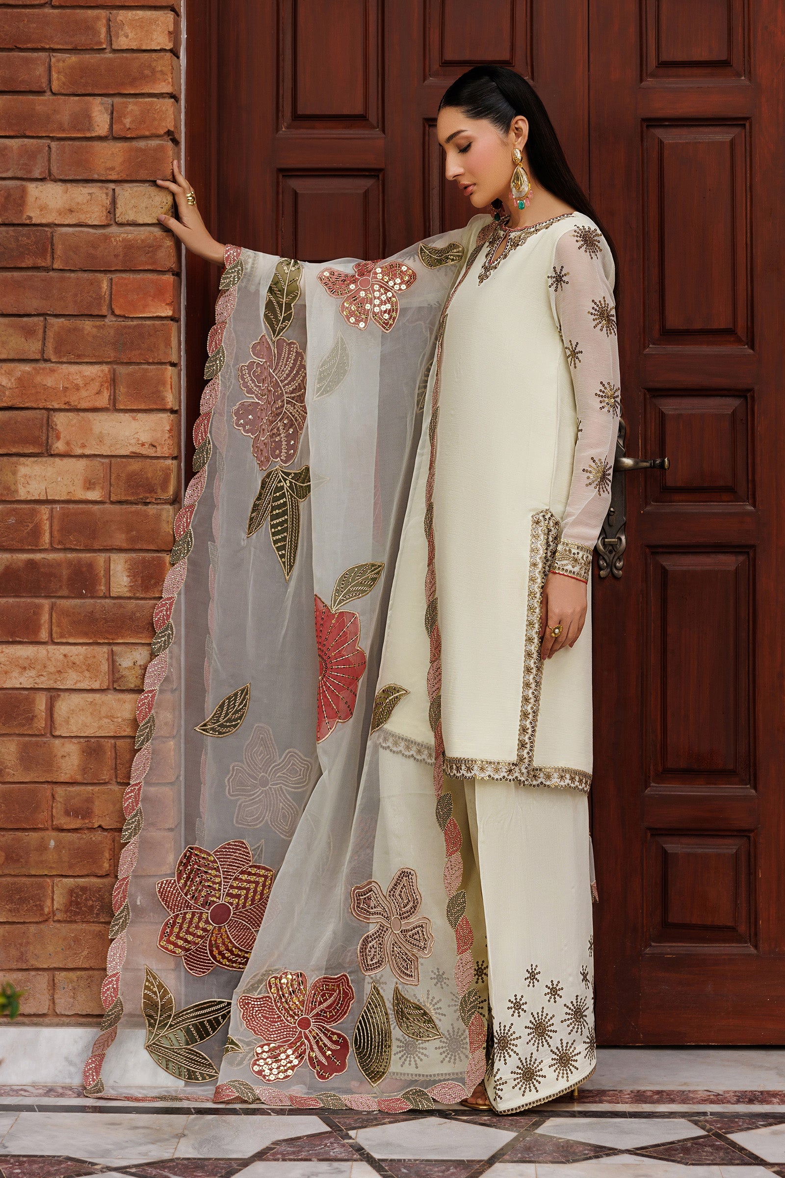 Woman in traditional outfit with floral dupatta standing against a brick wall and wooden door.