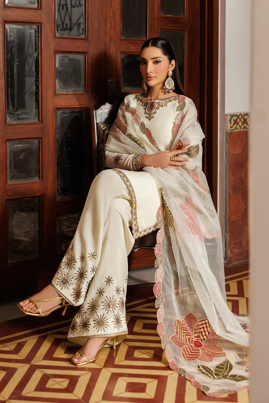 Woman in a traditional outfit with floral patterns sitting in a room with wooden walls and a patterned floor.