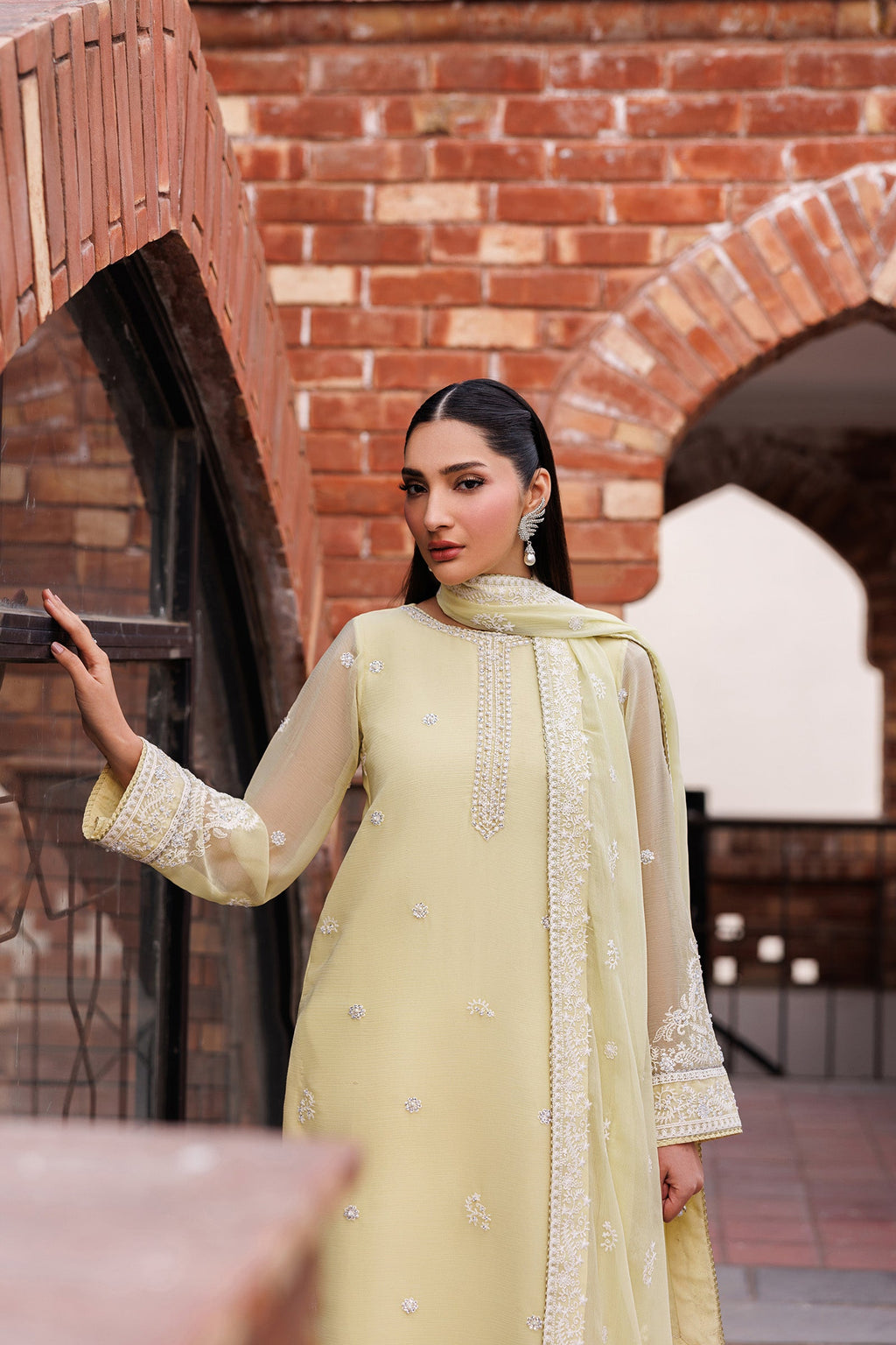 Woman in a light green traditional outfit with intricate patterns standing against a brick wall.