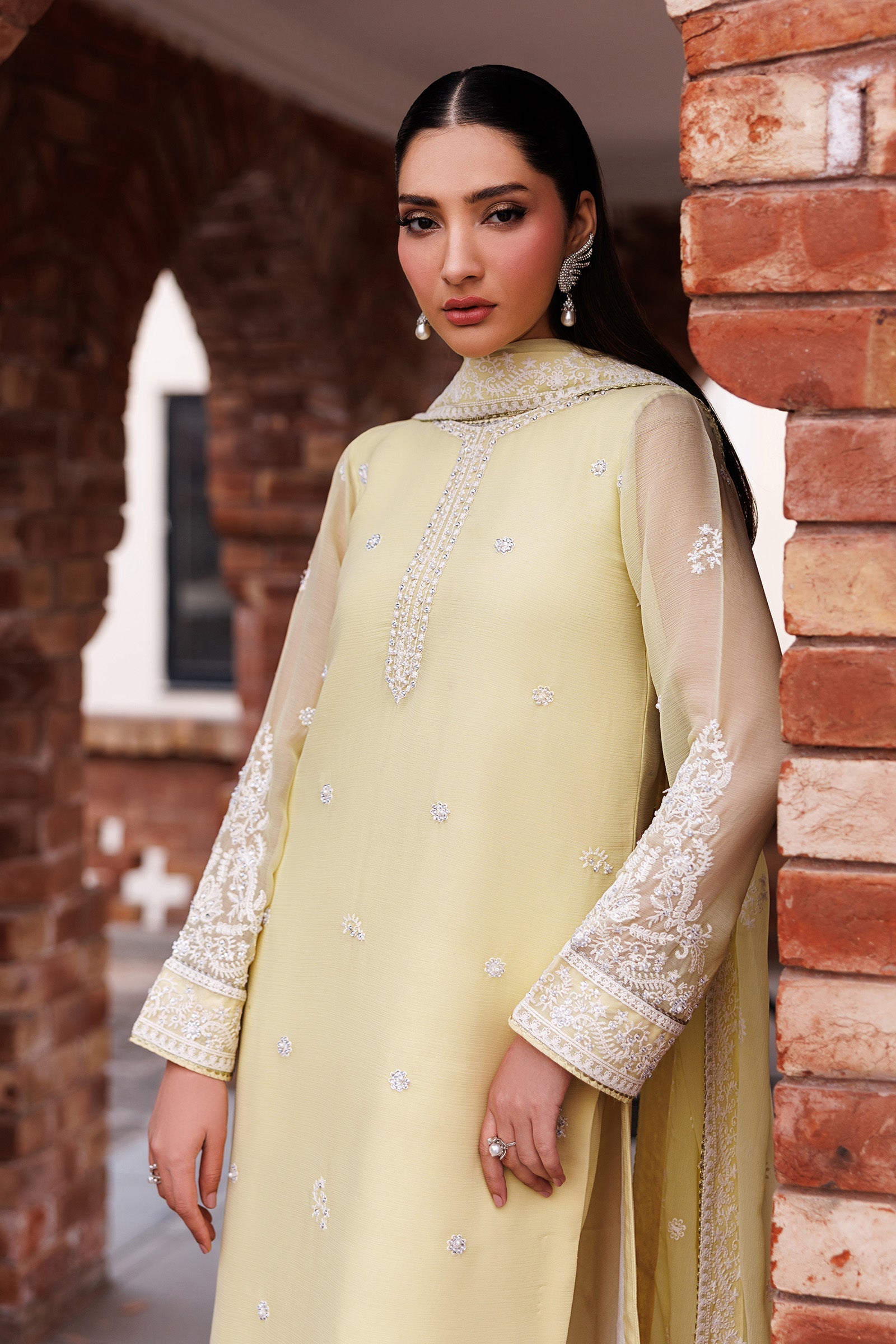 Woman in a light green traditional outfit with white embroidery standing against a brick wall.