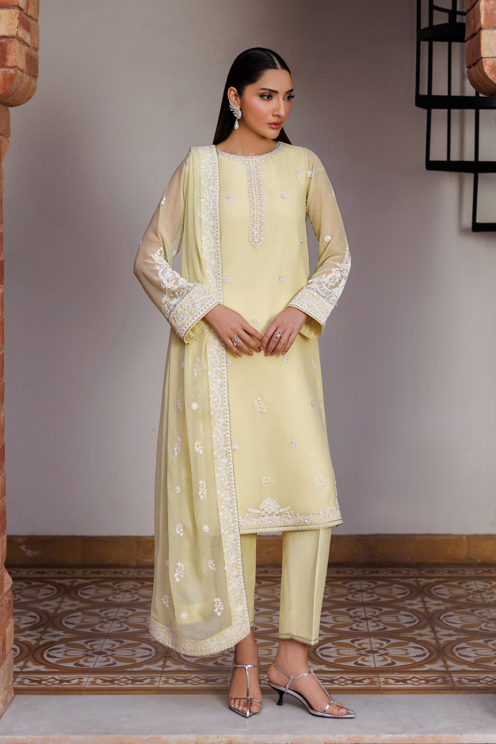 Woman wearing a light yellow traditional outfit with intricate patterns in a room with a patterned floor.