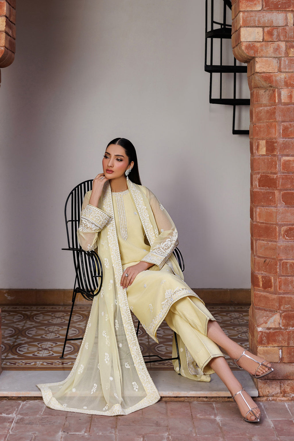Woman in a light yellow traditional outfit sitting on a chair against a brick wall.