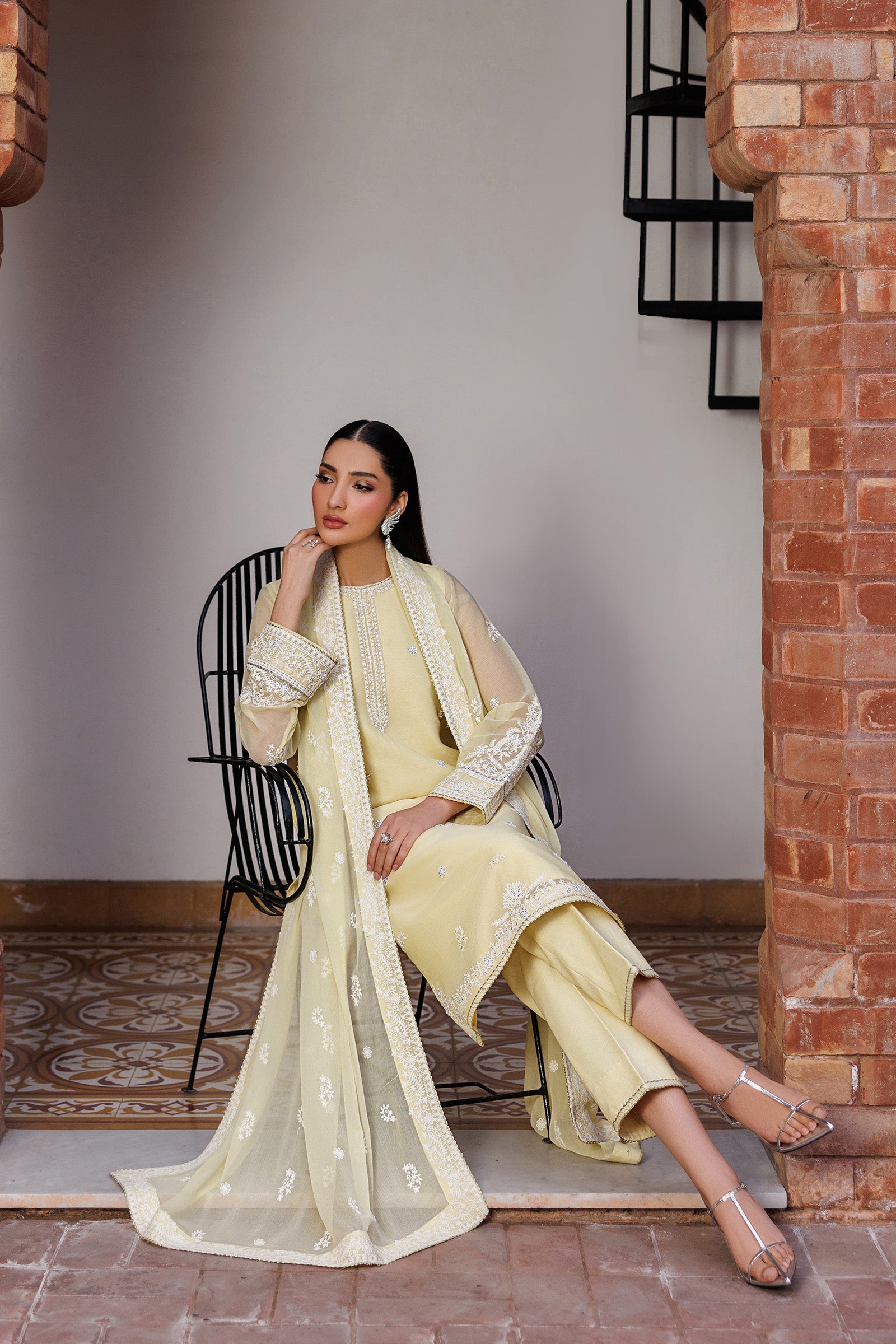 Woman in a light yellow traditional outfit sitting on a chair against a brick wall.