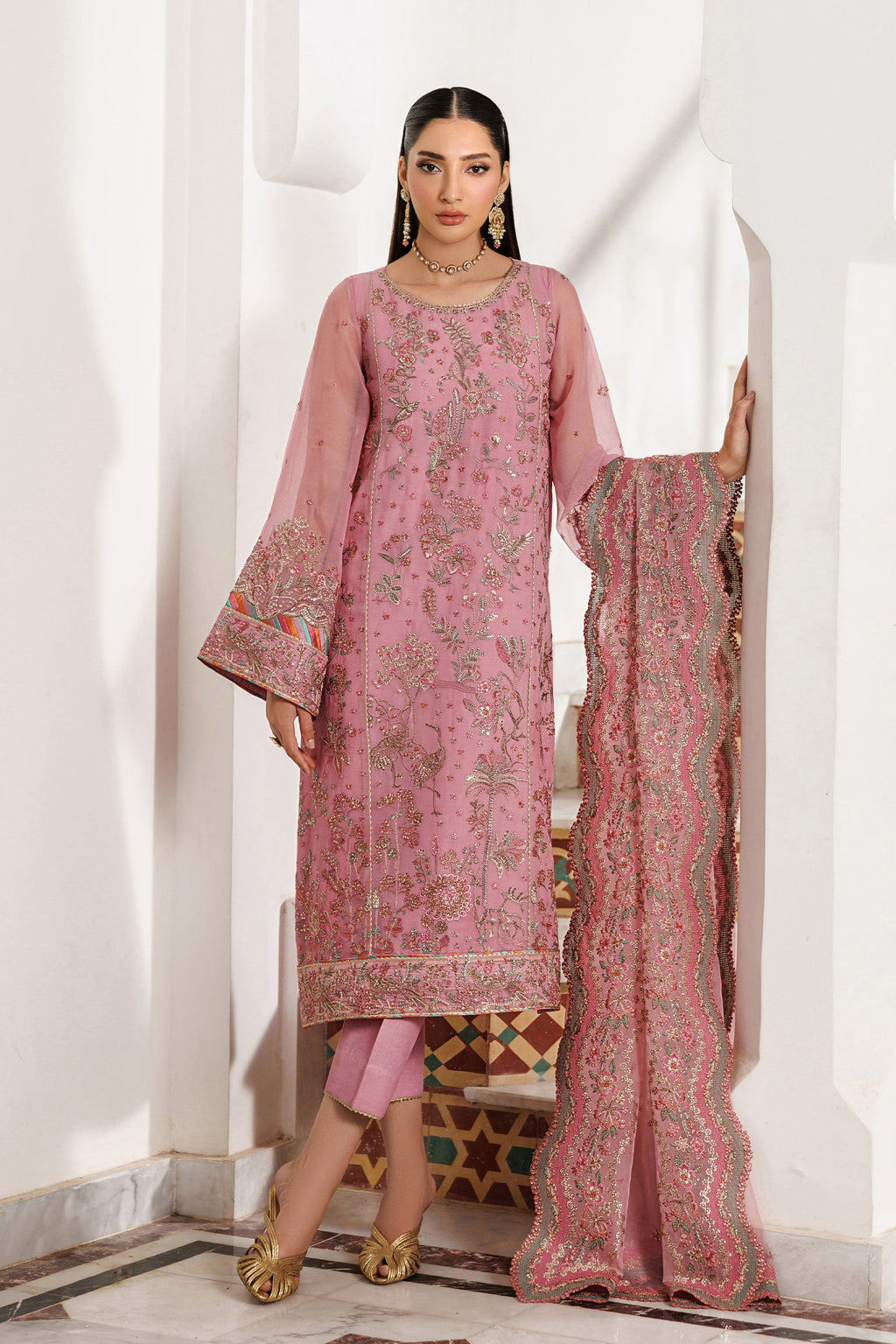 Woman wearing a pink embroidered traditional outfit with a matching dupatta against a white wall.