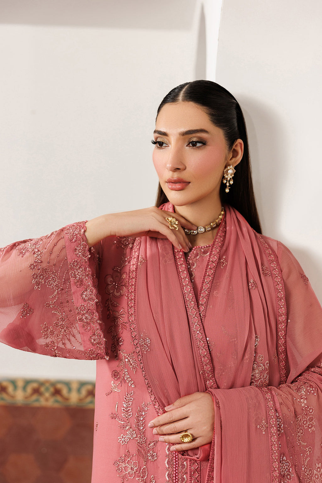 Woman wearing a pink embroidered traditional outfit against a white wall.