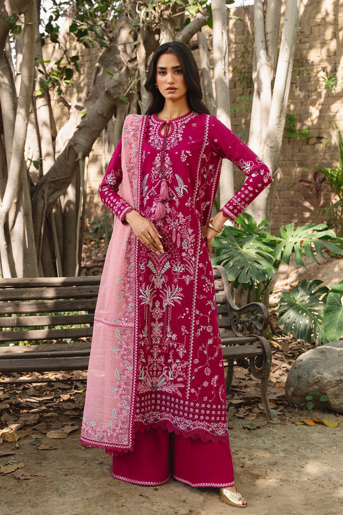Woman in a pink embroidered traditional outfit standing outdoors with trees and plants in the background