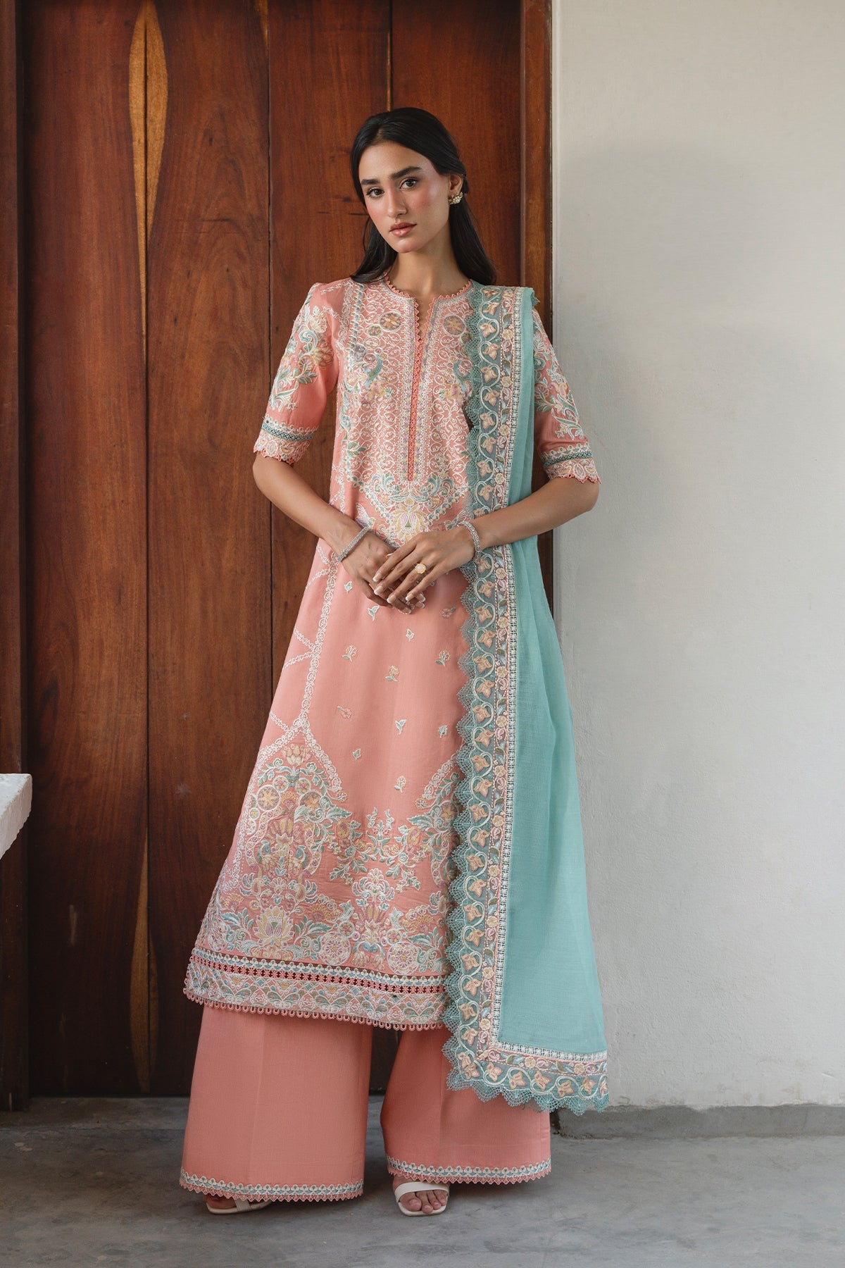 Woman wearing a traditional outfit with a pink and blue color scheme against a wooden and white wall.