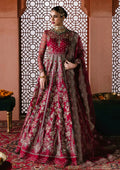 A woman models an ornate magenta and gold embroidered traditional gown with a matching sheer dupatta in an indoor setting with patterned screens and warm-toned walls.