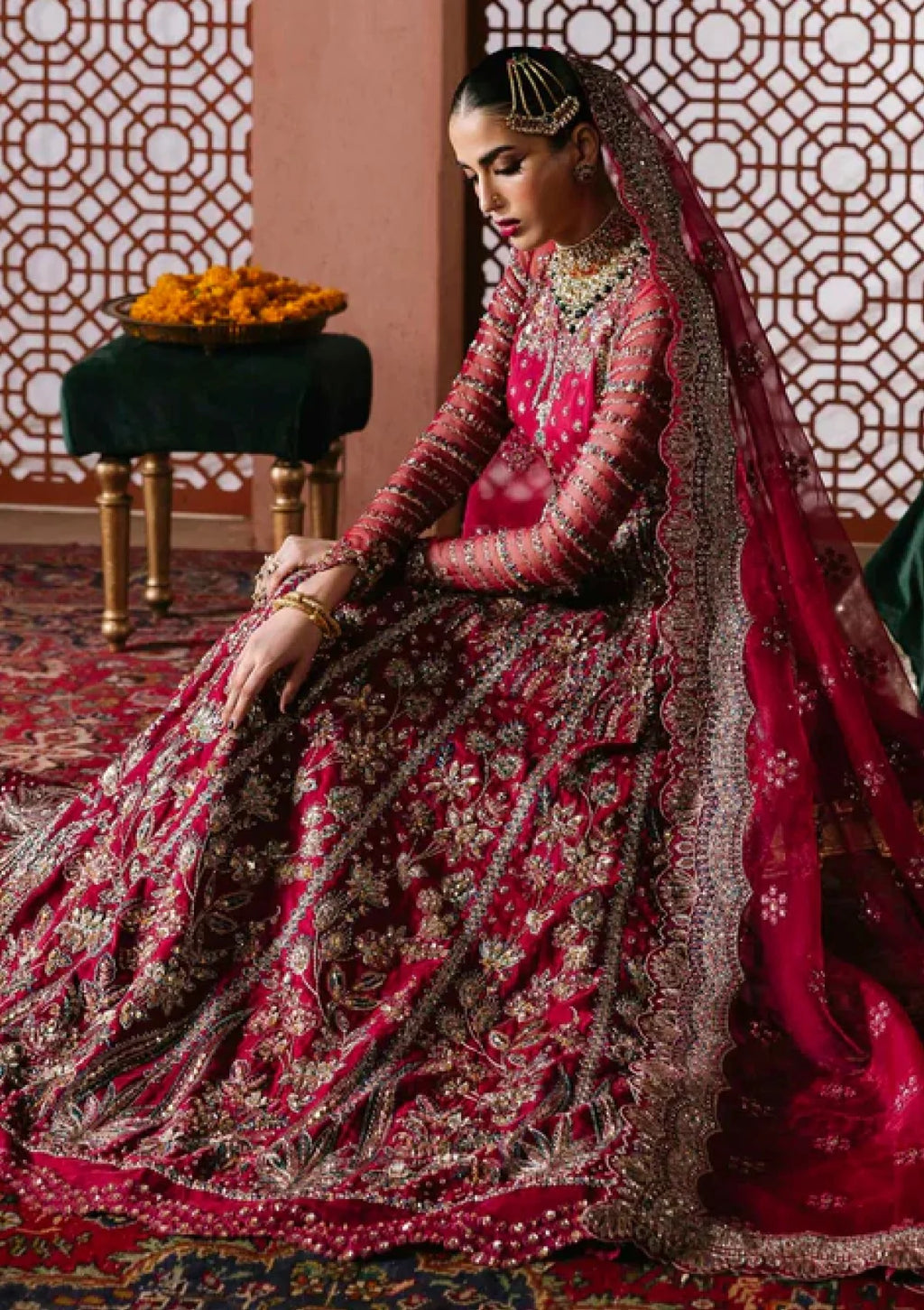 A woman seated on a patterned rug wearing an ornate magenta and gold embroidered traditional bridal outfit with a long veil, jewelry, and a decorative interior with geometric screens and a small table holding orange flowers.