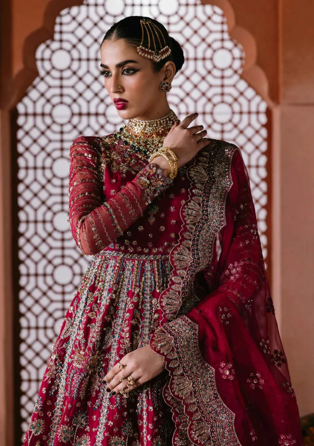 A woman models a traditional red and gold heavily embroidered Anarkali gown with a matching dupatta, set against an arched architectural background with a geometric screen.