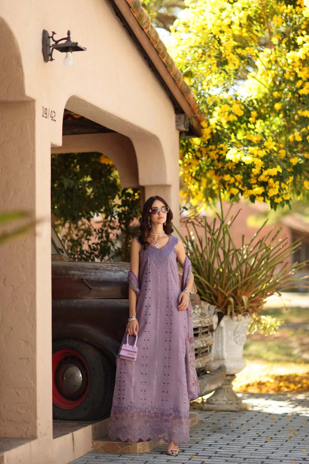 Woman in a purple dress standing in front of a vintage car with a yellow flowering tree in the background.