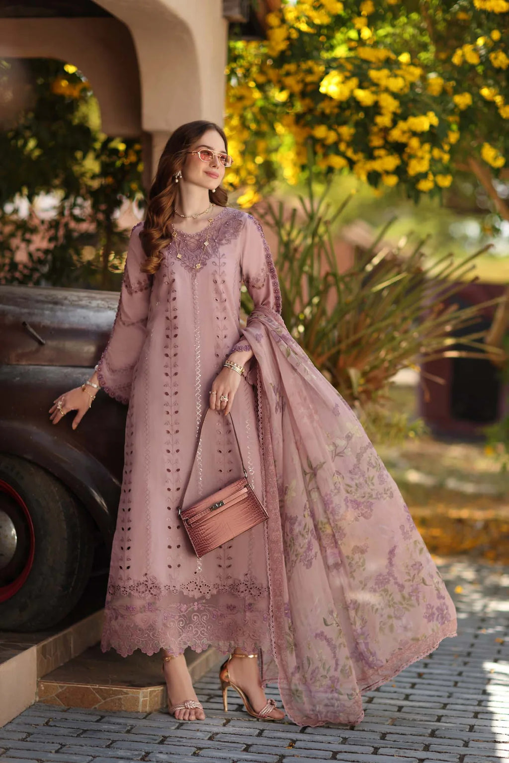 Woman in a pink traditional outfit standing next to a vintage car with yellow flowers in the background.