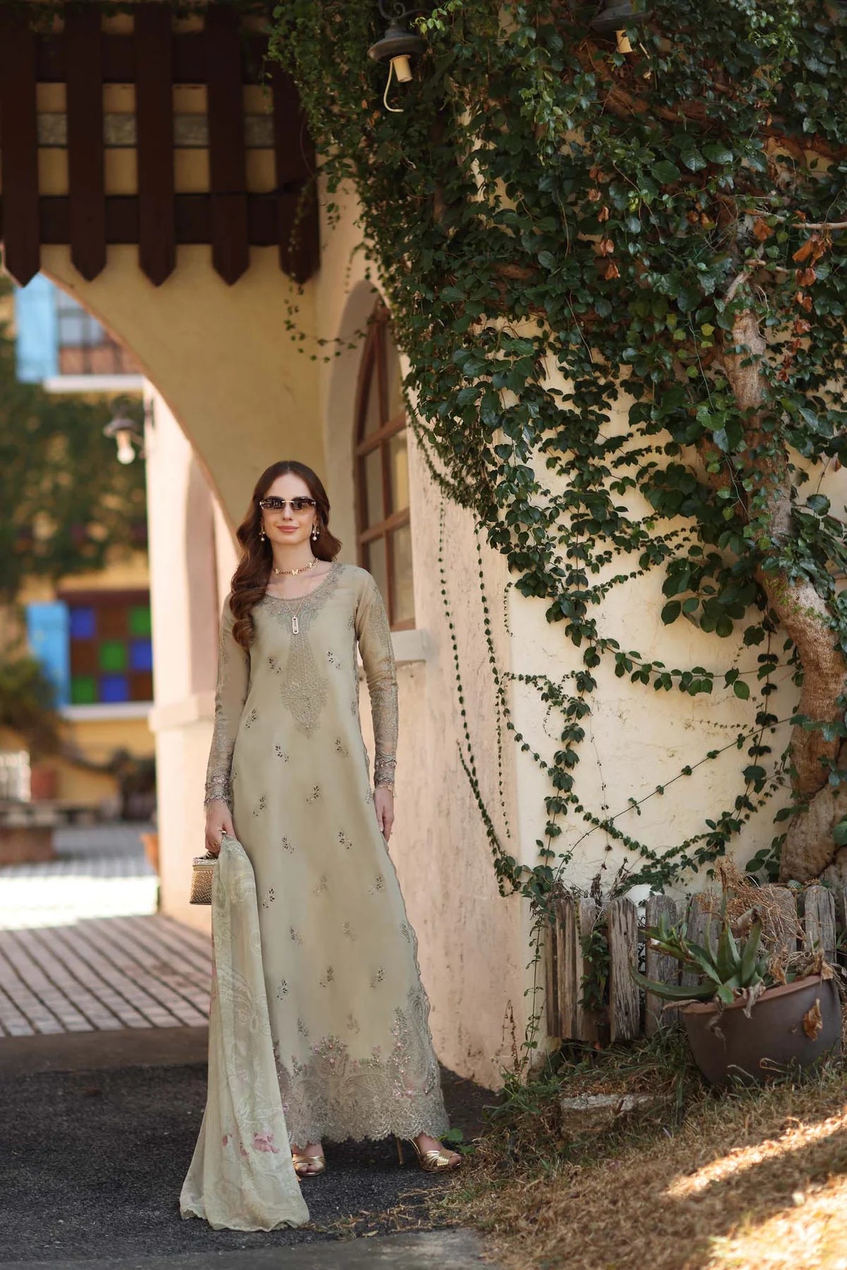 Woman in a long, light-colored dress standing outdoors with greenery and a building in the background.