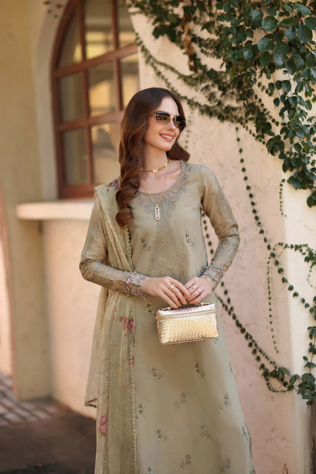 Woman in a light green traditional outfit holding a clutch, standing outdoors with decorative plants.