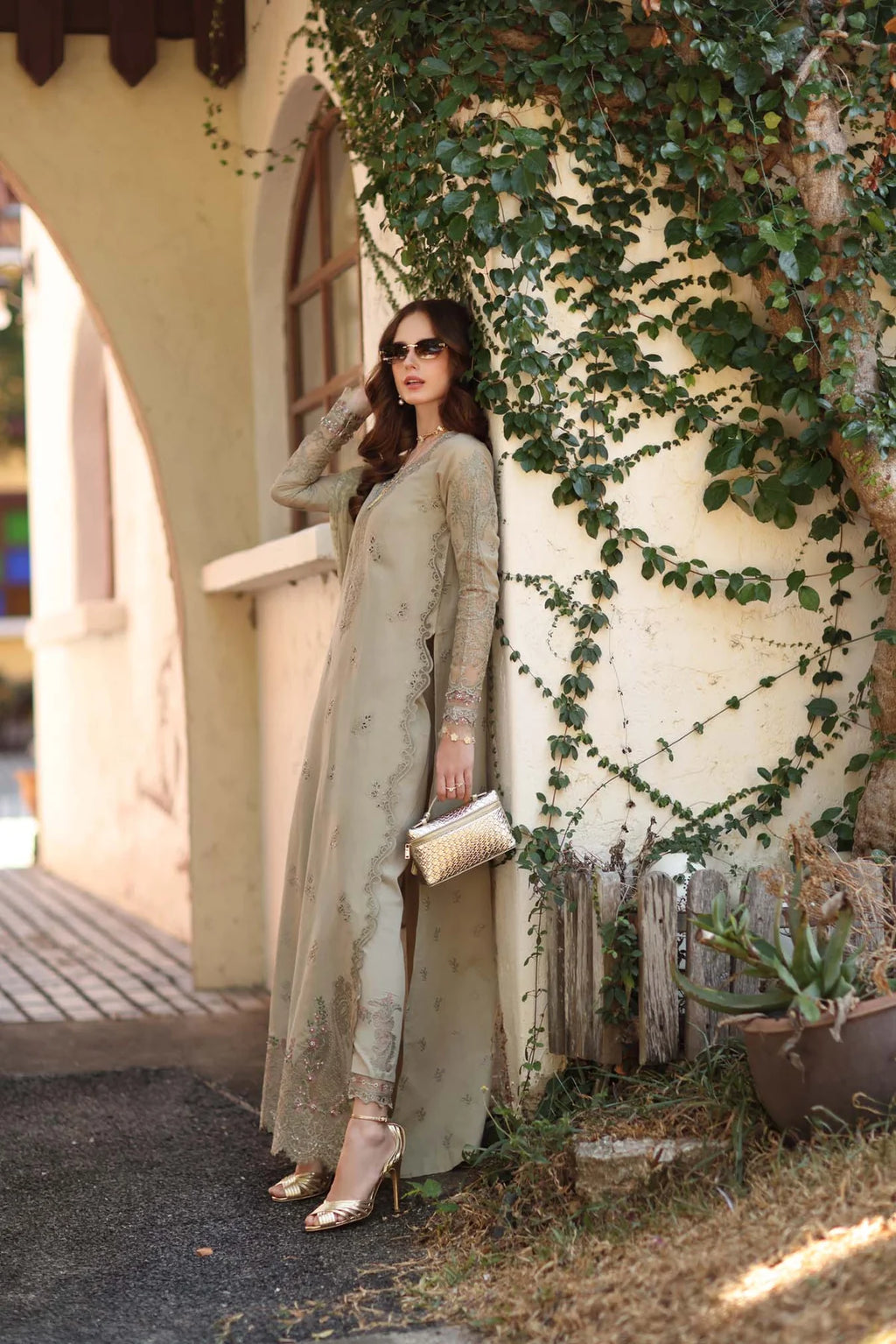 Woman in a long, light-colored dress standing against a wall with greenery