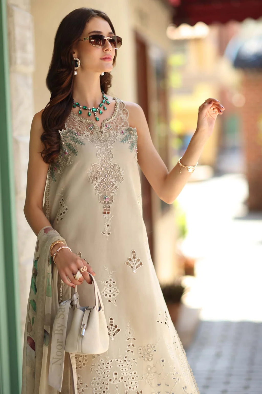 A woman wearing a cream-colored embroidered ethnic dress with intricate floral patterns, accessorized with a matching scarf, sunglasses, and jewelry, standing in a sunlit outdoor setting.