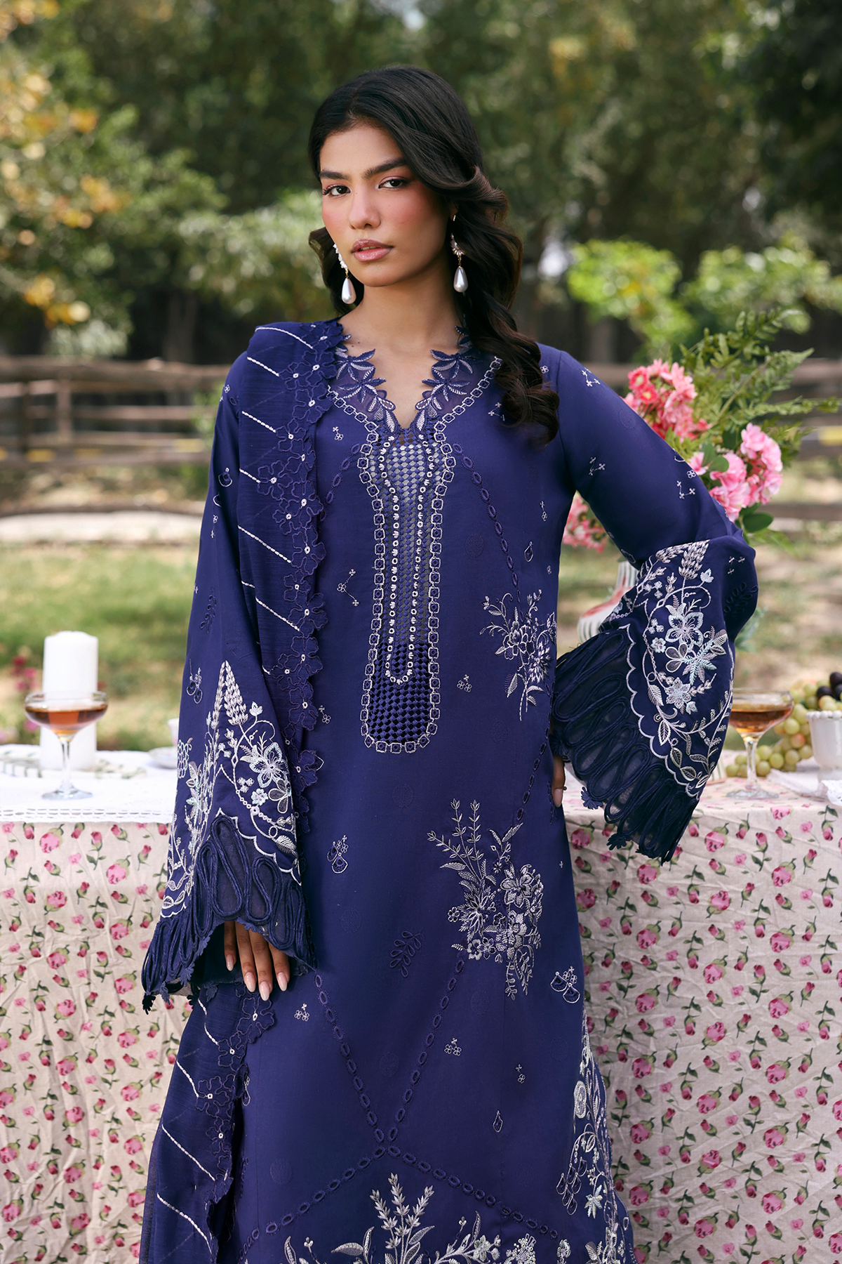 Woman in a blue embroidered dress standing outdoors at a table with flowers and candles.