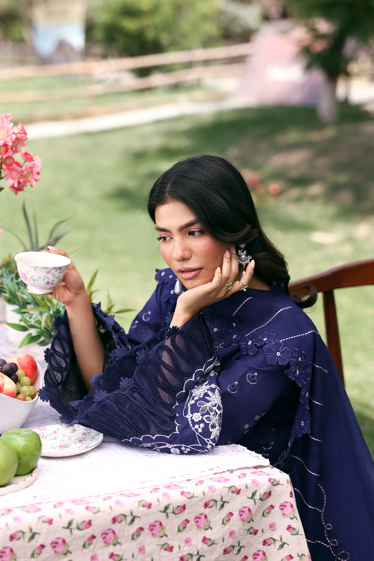 Woman in a blue embroidered dress sitting at a table outdoors, holding a teacup.