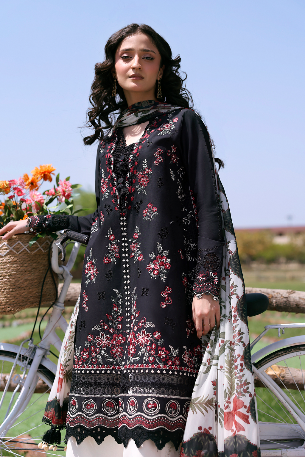 Woman wearing a black floral dress with a scarf, standing outdoors with a bicycle and flowers in the background.