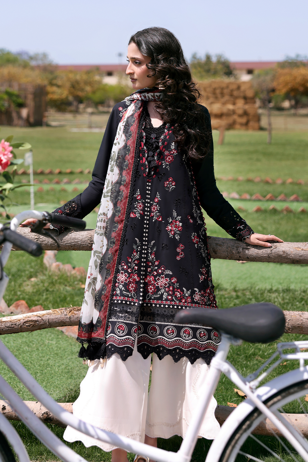 Woman in traditional black and white floral outfit standing next to a bicycle on a grassy field.