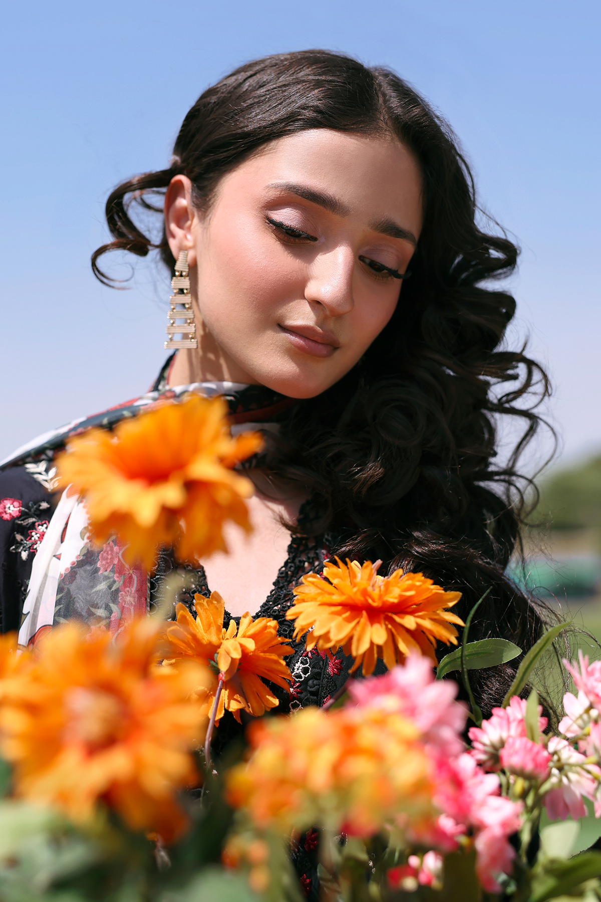 Woman with dark hair and earrings surrounded by orange and pink flowers outdoors.