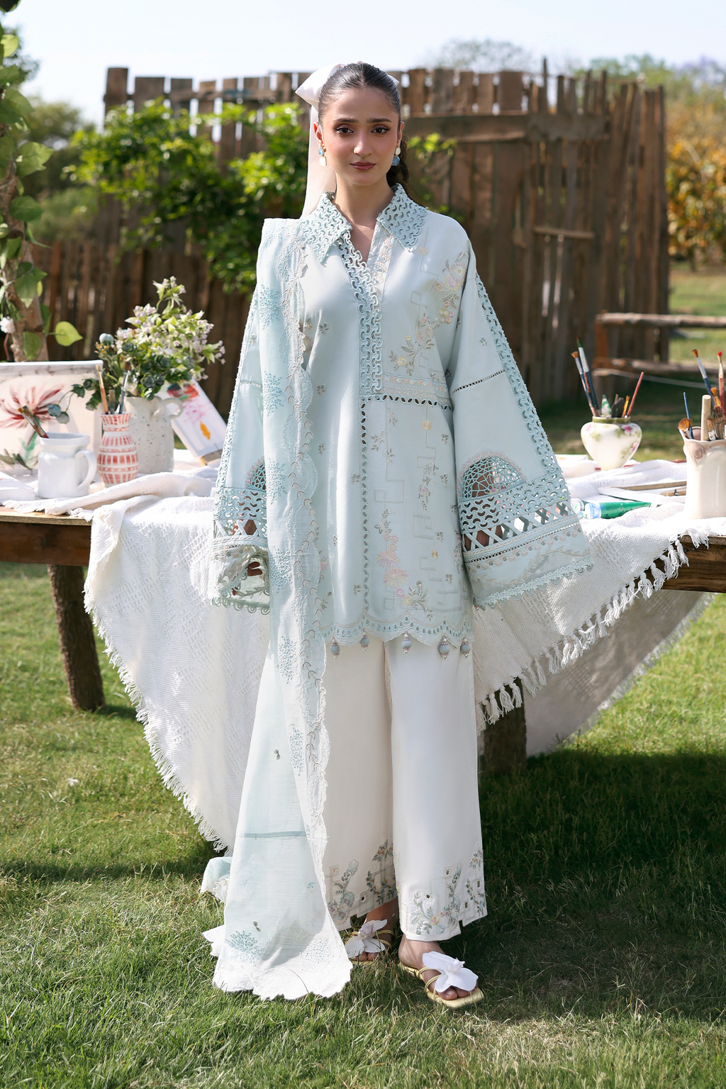 Woman in a light blue embroidered outfit standing outdoors with a table and plants in the background